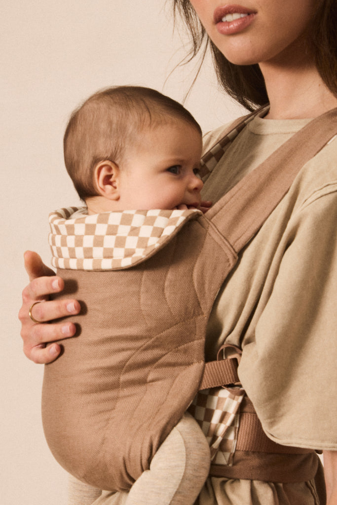A close-up lifestyle shot of a baby in a tan quilted front baby carrier worn by a mother. The carrier features a beige and white checkered fabric lining on the neck and waist straps, set against a warm beige background.