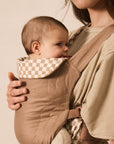 A close-up lifestyle shot of a baby in a tan quilted front baby carrier worn by a mother. The carrier features a beige and white checkered fabric lining on the neck and waist straps, set against a warm beige background.