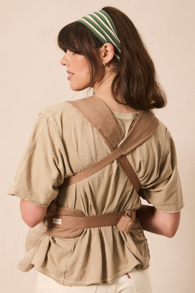 A woman shown from the back wearing a beige baby carrier wrap styled over a matching beige short-sleeve top, with a green and white striped headband, against a solid beige background.