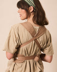 A woman shown from the back wearing a beige baby carrier wrap styled over a matching beige short-sleeve top, with a green and white striped headband, against a solid beige background.