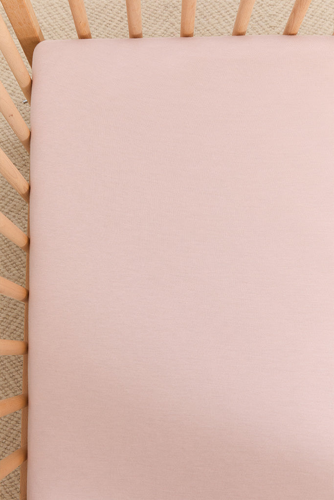 Light pink fitted crib sheet on a wooden crib mattress with beige carpet visible underneath.