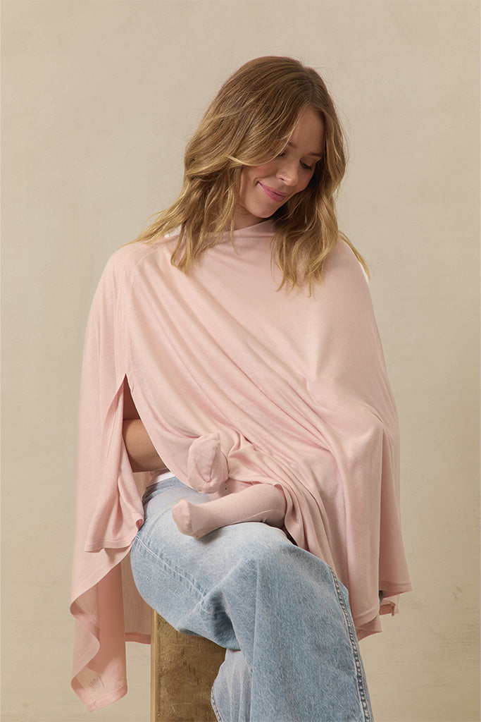 Woman wearing a light pink nursing cover while breastfeeding a baby in light pink socks, sitting on a wooden stool.