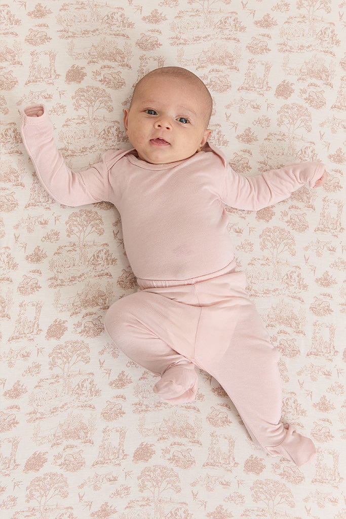 Baby wearing solid light pink long-sleeve footed pajamas lying on beige patterned blanket.