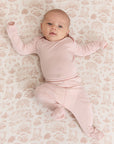 Baby wearing solid light pink long-sleeve footed pajamas lying on beige patterned blanket.