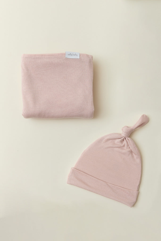 Soft pink baby swaddle blanket and matching knotted hat set on a cream background.