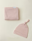 Soft pink baby swaddle blanket and matching knotted hat set on a cream background.