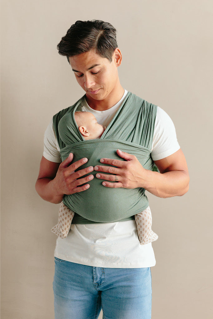 Man wearing a white shirt and blue jeans using a green fabric baby carrier holding a sleeping infant.