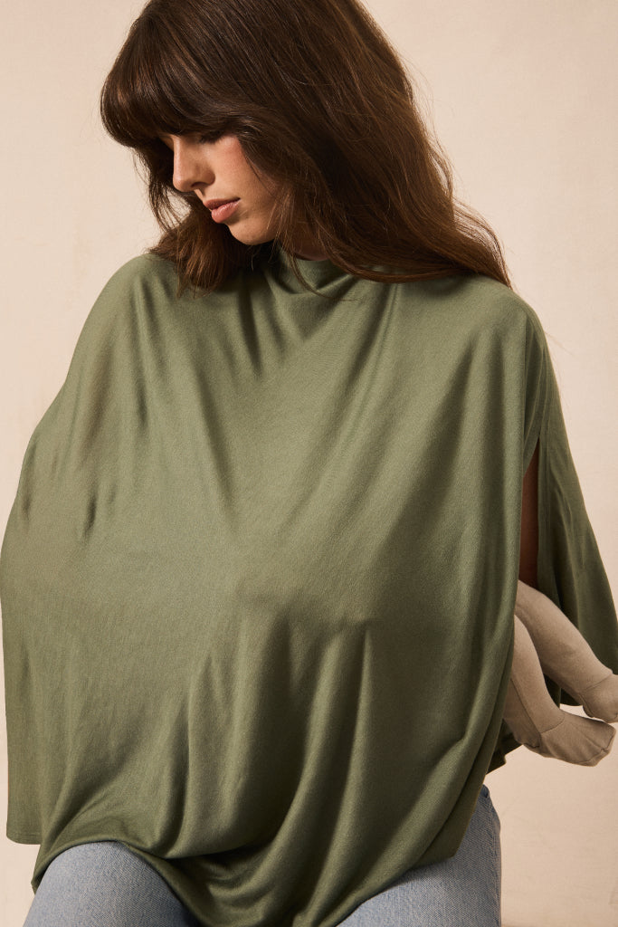 A woman wearing an oversized olive green mock-neck top or poncho-style sweater, paired with light blue jeans, posed against a plain beige background.
