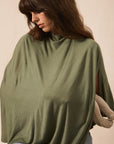 A woman wearing an oversized olive green mock-neck top or poncho-style sweater, paired with light blue jeans, posed against a plain beige background.