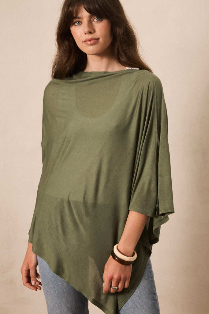 A woman wearing an olive green draped, semi-sheer long-sleeve top with an asymmetrical hem, paired with light blue jeans, against a plain beige background.
