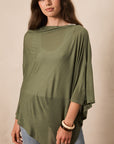 A woman wearing an olive green draped, semi-sheer long-sleeve top with an asymmetrical hem, paired with light blue jeans, against a plain beige background.