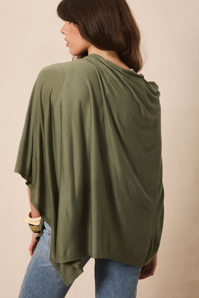 A woman wearing an olive green draped poncho-style top with blue jeans, shown from the back against a plain beige studio background.