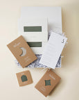Gift box set with olive green baby sleeper, hat, and swaddle in kraft paper packaging, plus product information card.