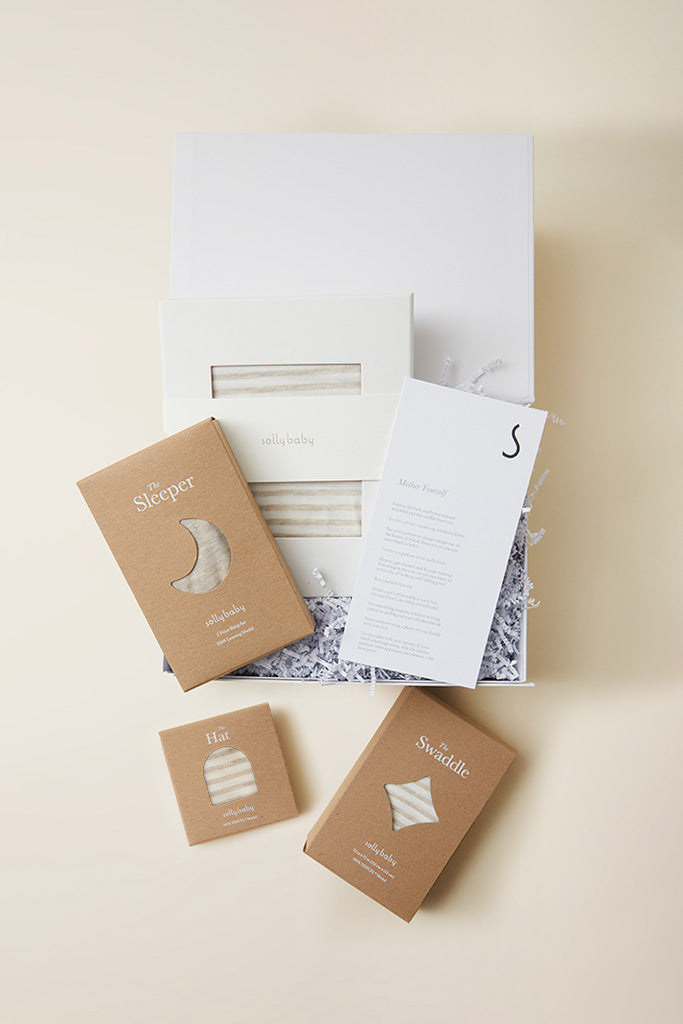 Baby gift set with beige striped sleeper, swaddle, and hat in brown packaging, displayed in white box with shredded paper.