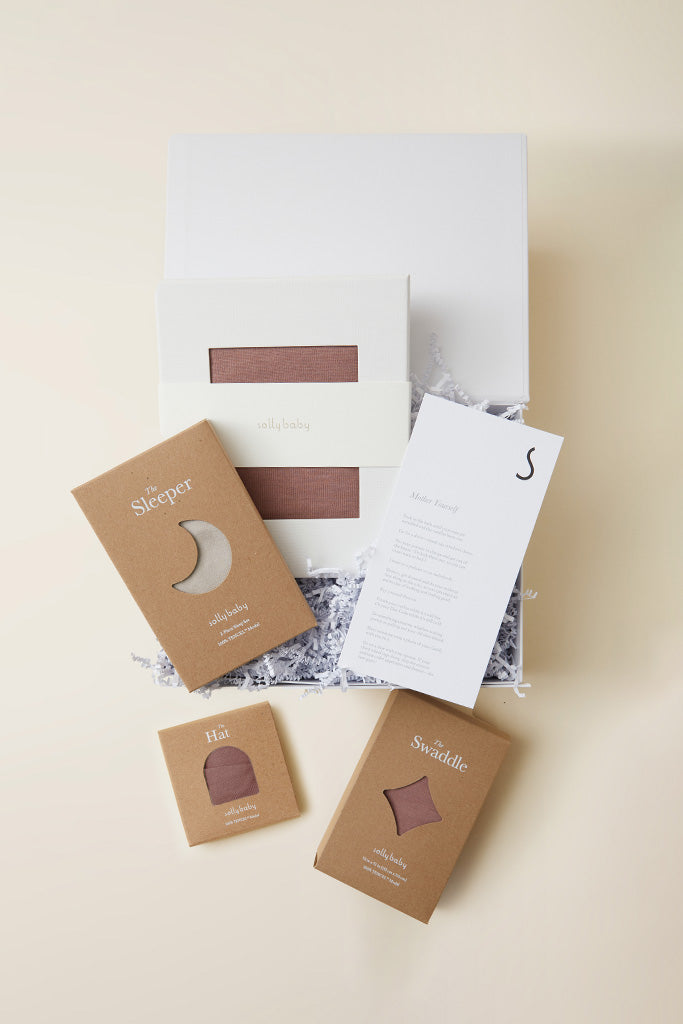 Brown baby swaddle, sleeper, and hat packaged in cardboard boxes with white shredded filler in an open white gift box.