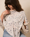A woman wearing a cream-colored wrap top with a blue floral pattern and light blue jeans, posed against a neutral beige background.