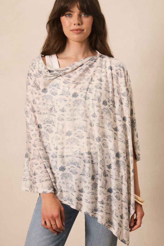A woman wearing a draped, lightweight poncho-style top with a blue floral toile pattern on a cream background, paired with light-wash blue jeans, set against a plain beige studio background.