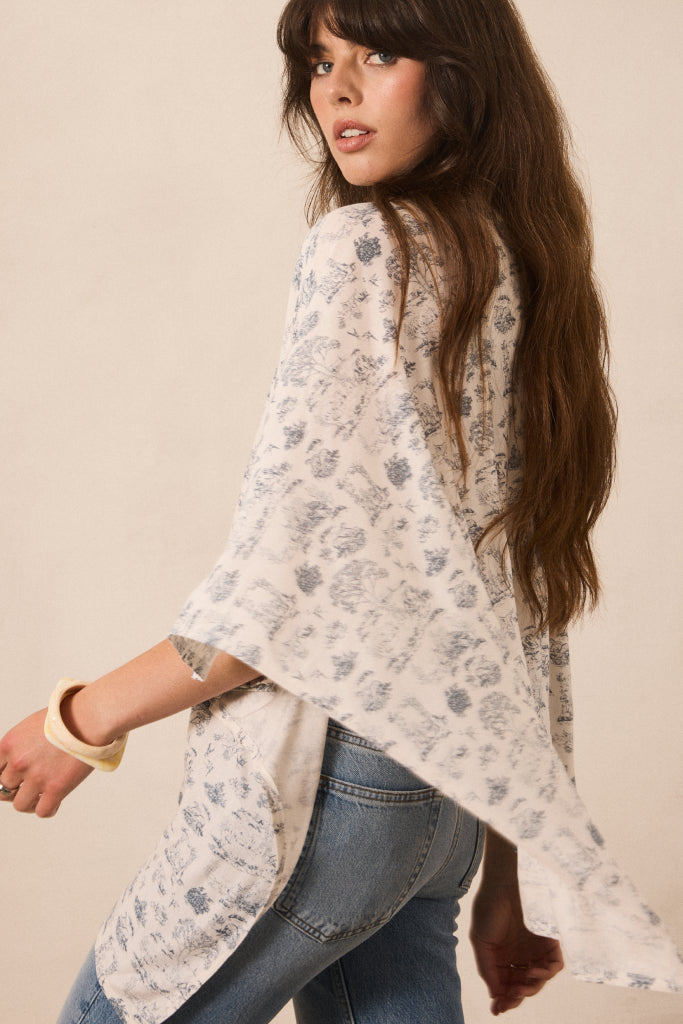 A woman wearing a loose, off-white floral kimono-style top with blue jeans, photographed against a plain beige studio background.