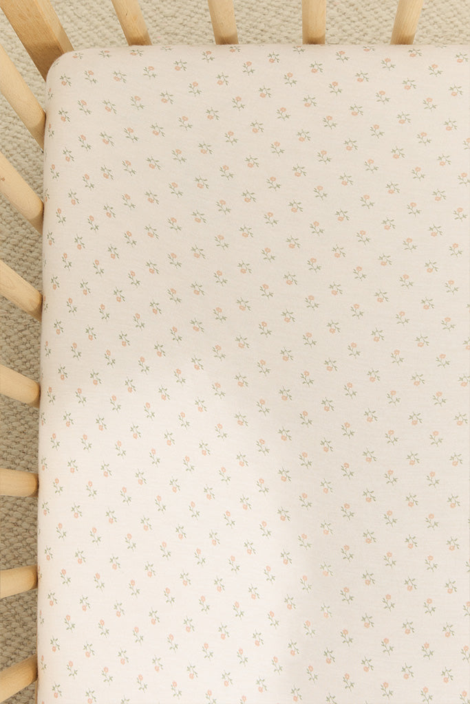 Crib mattress covered with white fitted sheet featuring small pink floral pattern inside light wooden crib.