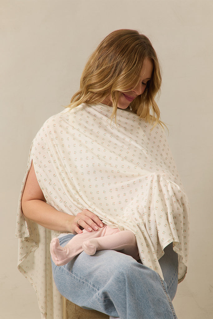 Cream nursing cover with small floral print worn by a woman breastfeeding a baby in pink tights and light denim jeans