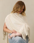 Cream nursing cover with small floral print worn by a woman breastfeeding a baby in pink tights and light denim jeans