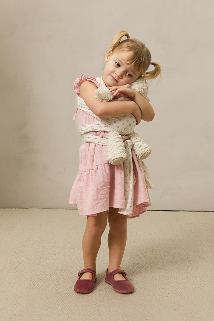 Toddler girl in pink dress and maroon shoes hugging cream plush toy secured with floral wrap carrier.