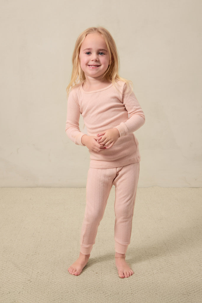 Young girl wearing a light pink long-sleeve ribbed top and matching ribbed pants standing barefoot.