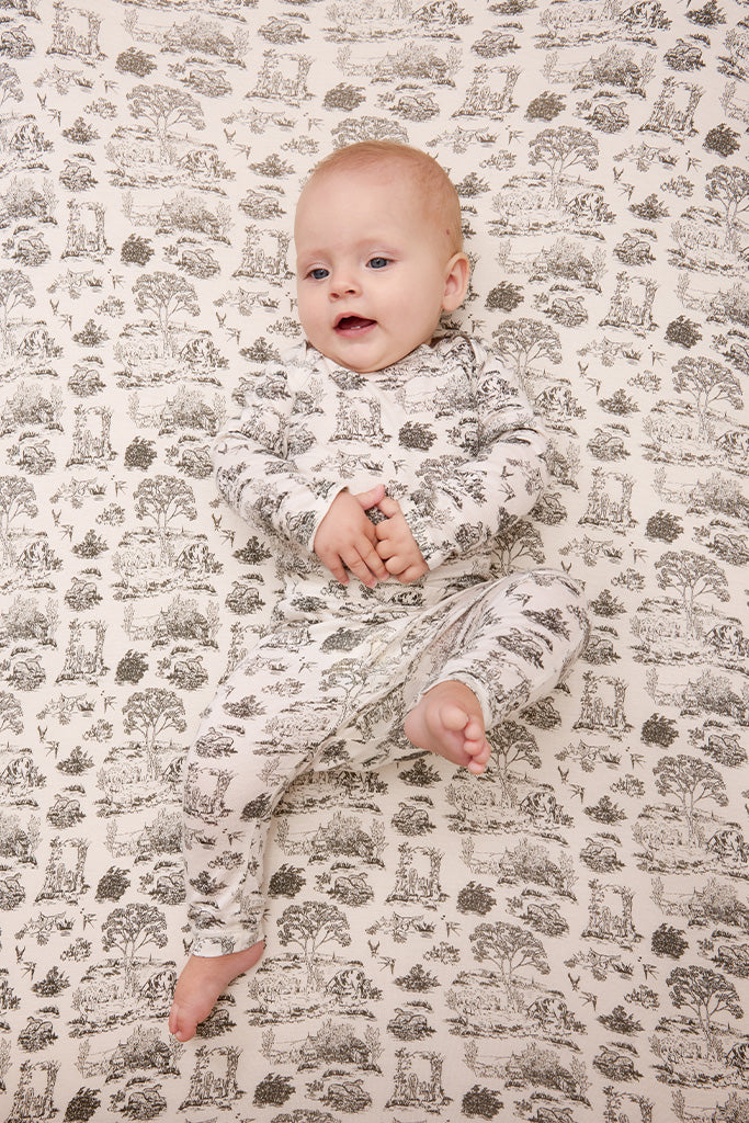 Baby wearing long-sleeve cream sleeper with brown nature scenes print, lying on matching printed blanket.