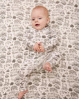 Baby wearing long-sleeve cream sleeper with brown nature scenes print, lying on matching printed blanket.