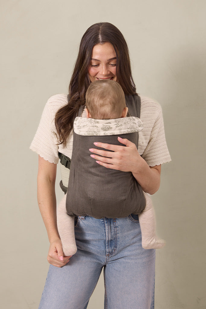 Woman wearing a dark gray baby carrier with beige patterned head support holding a baby facing inward.