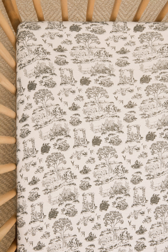 Cream fitted crib sheet with black toile pattern featuring pastoral scenes and animals in wooden crib.