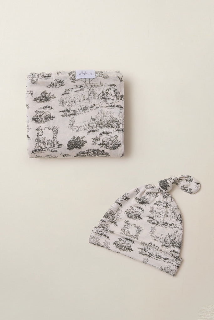 Folded beige baby blanket and matching knotted baby hat with black countryside animal and tree print pattern.