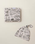 Folded beige baby blanket and matching knotted baby hat with black countryside animal and tree print pattern.