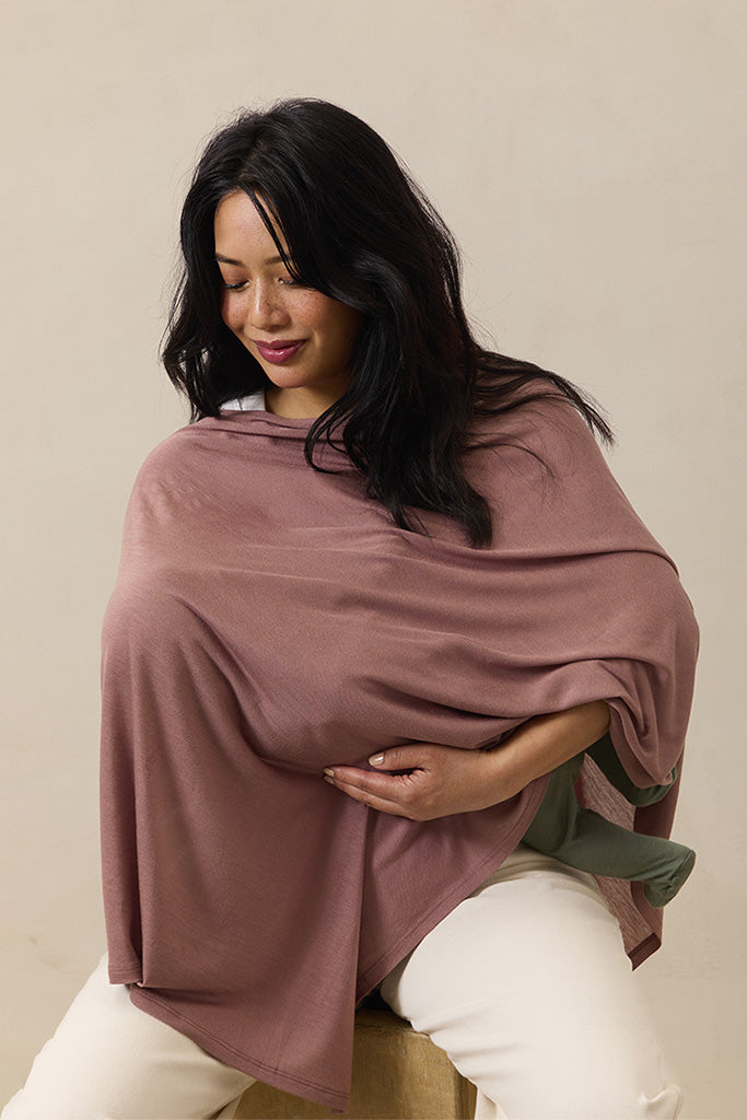 Woman wearing dusty rose nursing cover while holding a baby dressed in olive green pants and white top