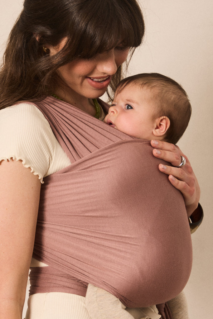 A woman wearing a dusty rose baby wrap carrier with a baby inside, photographed against a plain beige background.