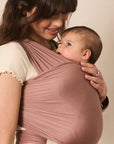 A woman wearing a dusty rose baby wrap carrier with a baby inside, photographed against a plain beige background.