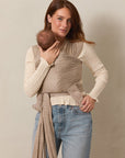 A woman wearing a white blouse and jeans standing and holding a baby in a Solly Baby Wrap carrier in the Cream Floral print, featuring delicate cream flowers on a taupe background. 