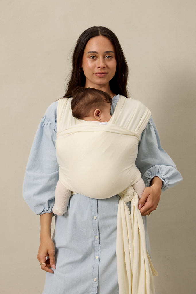 Woman wearing a baby in a cream pointelle solly baby wrap against a beige background