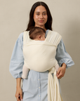 Woman wearing a baby in a cream pointelle solly baby wrap against a beige background