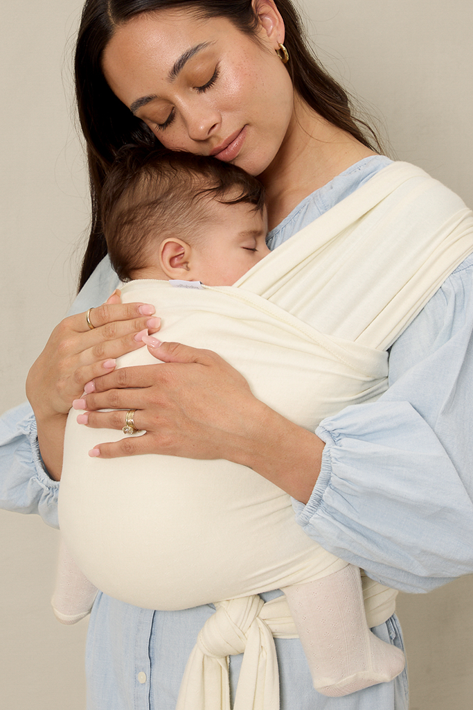 Woman holding a baby in a cream pointelle solly baby wrap against a neutral background
