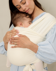 Woman holding a baby in a cream pointelle solly baby wrap against a neutral background