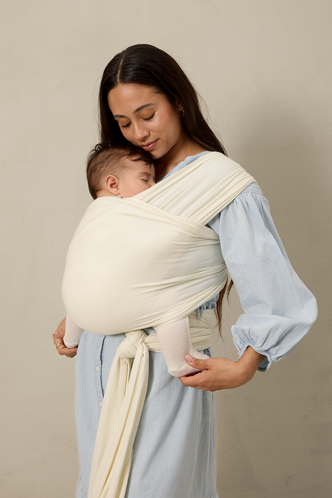 Woman holding a baby in a cream pointelle solly baby wrap against a beige background