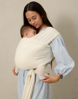 Woman holding a baby in a cream pointelle solly baby wrap against a beige background
