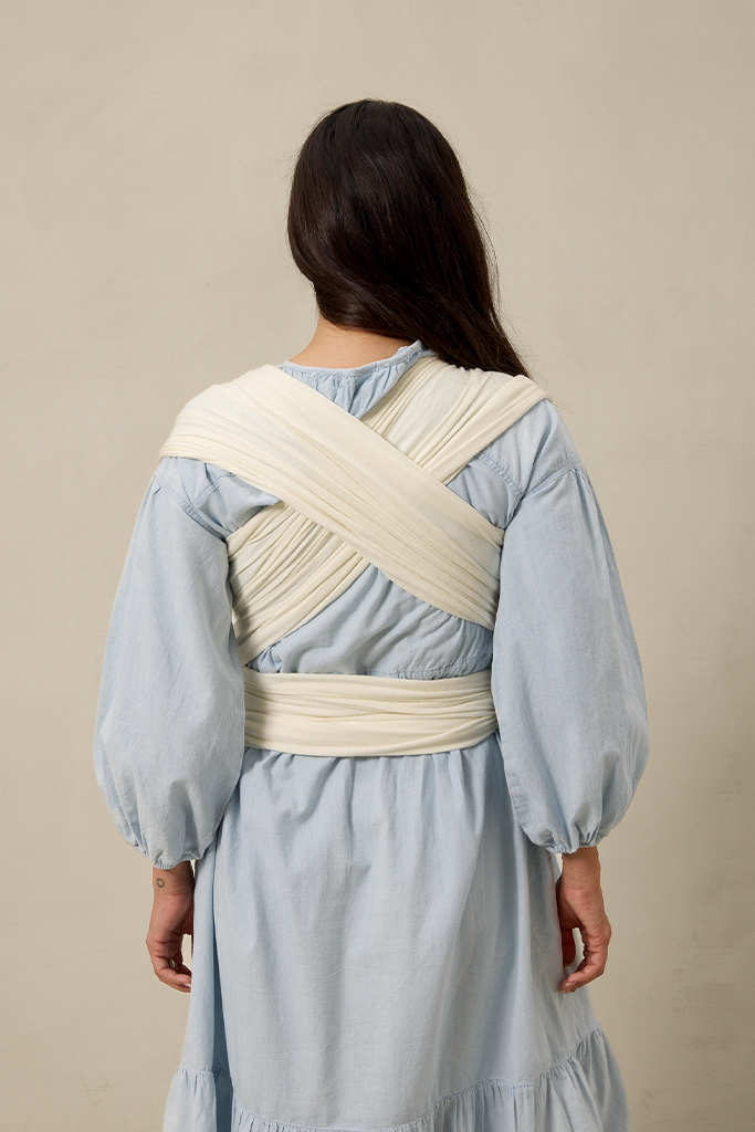 Woman wearing a light blue dress with a cream pointelle solly baby wrap around her shoulders against a beige background