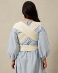 Woman wearing a light blue dress with a cream pointelle solly baby wrap around her shoulders against a beige background