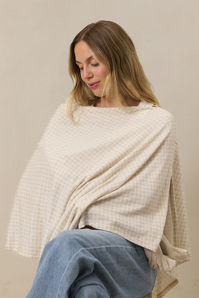 Woman wearing beige houndstooth patterned nursing cover over white top and light blue jeans against neutral background