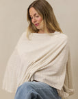 Woman wearing beige houndstooth patterned nursing cover over white top and light blue jeans against neutral background