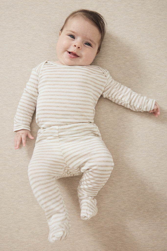 A smiling baby lying on their back wearing a cream and beige horizontally striped long-sleeve footed sleeper, set against a neutral beige textured background.