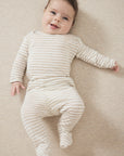 A smiling baby lying on their back wearing a cream and beige horizontally striped long-sleeve footed sleeper, set against a neutral beige textured background.