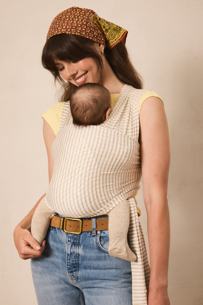 A woman wearing a cream and beige striped baby carrier wrap with a matching headscarf, carrying a baby against her chest. She is wearing a yellow top, blue jeans, and a brown belt, set against a plain beige studio background.